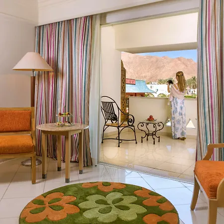Movenpick Taba Resort & Spa Exterior photo The image shows a cozy hotel room or apartment. It features a small living area with two orange chairs and a round table. The floor is decorated with a large circular green rug with colorful flowers. There are striped curtains framing a sliding door 