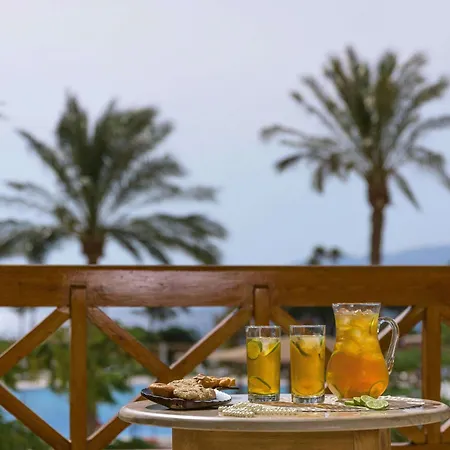Movenpick Taba Resort & Spa Exterior photo The photo shows a table set on a balcony with a view of palm trees in the background. On the table, there are three glasses filled with a yellow drink, likely iced tea or lemonade, garnished with lemon slices. Next to the glasses, there is a pitcher 