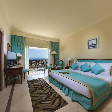 Movenpick Taba Resort & Spa Exterior photo The photo depicts a well-furnished hotel room featuring a large bed with light blue accents and decorative pillows. There are two lamps on either side of the bed, and a small table is positioned near the window, which is dressed with sheer curtains t